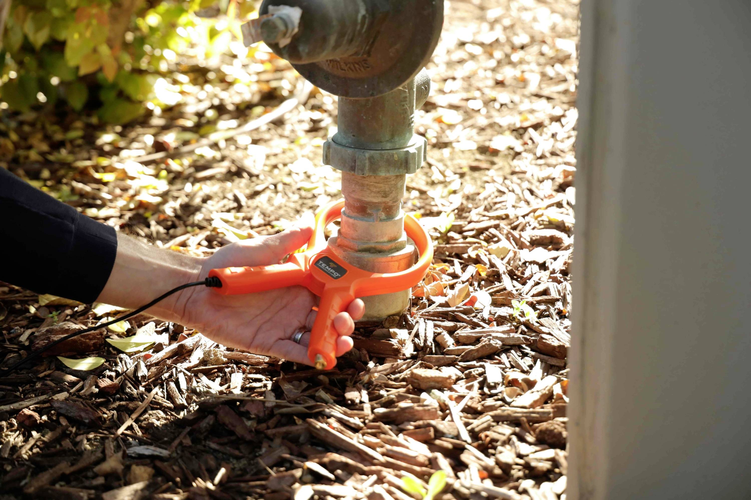 Een hand houdt een oranje waterlek detectieapparaat vast rond een buitenpijp boven een laag mulch, wat wijst op het uitvoeren van tests of onderhoudswerkzaamheden.