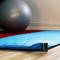 Een blauwe yogamat op een houten vloer met een grote grijze fitnessbal op de achtergrond en een kleine zwarte speaker aan de rand van de mat.