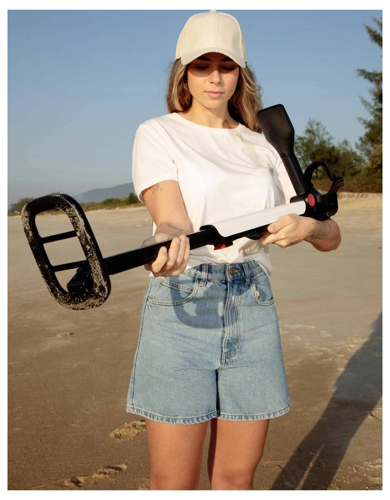 Een persoon in een wit shirt en spijkerstort staat op een strand, met een metaaldetector in de hand, wat suggereert dat hij op zoek is naar begraven voorwerpen.