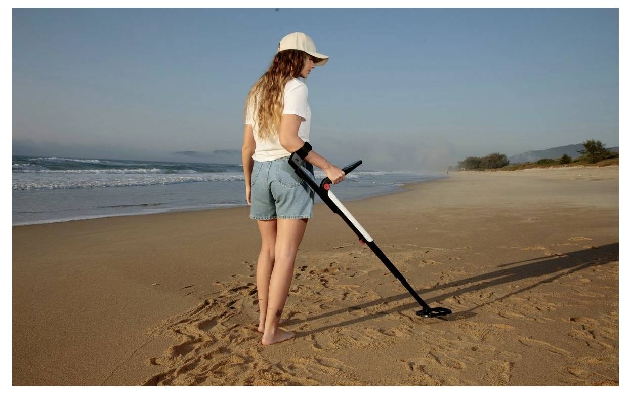 Een persoon met een pet gebruikt een metaaldetector op een leeg strand, terwijl hij het zand bij de waterlijn afscant bij zonsopgang.