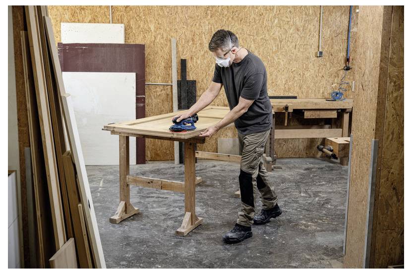 Een persoon met een masker staat een houten deur te schuren op een werkbank in een timmerwerkplaats vol verschillende houten materialen.