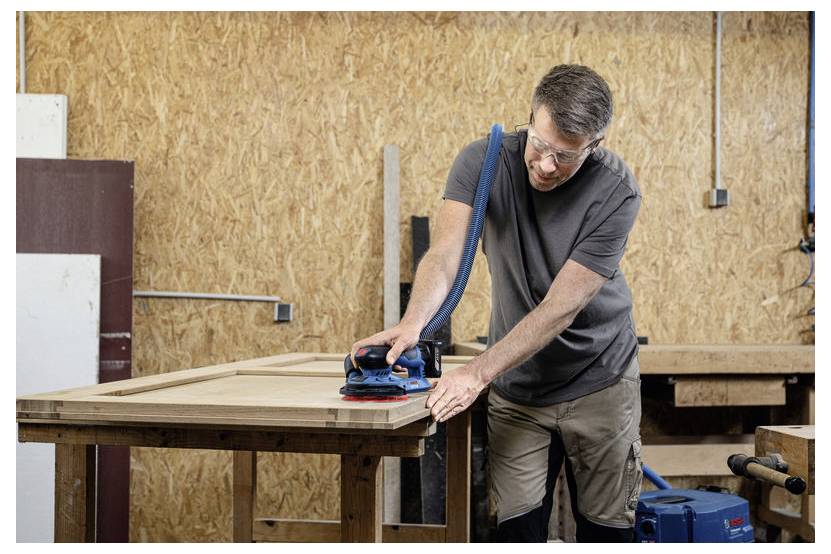 Een man met veiligheidsbril slijpt een houten plank in een werkplaats, waarbij hij een elektrische schuurmachine gebruikt die is aangesloten op een stofafzuiger.