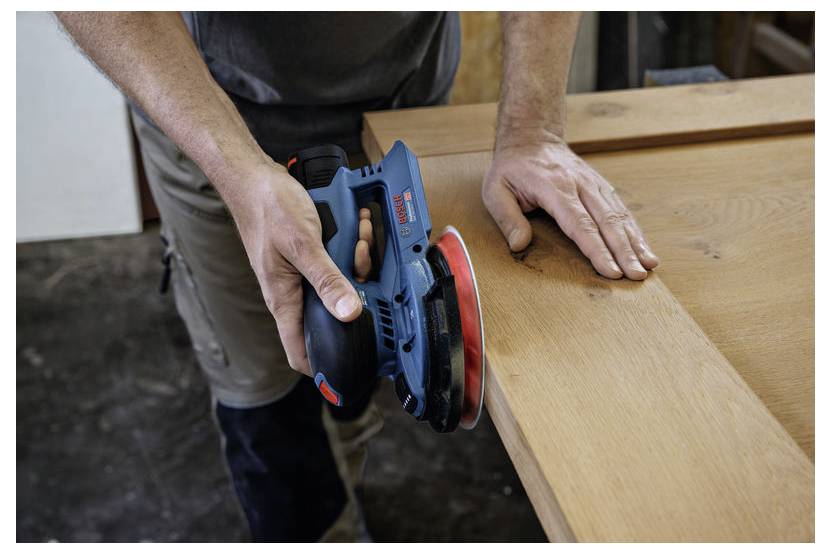 Een persoon gebruikt een elektrische schuurmachine op een houten oppervlak, waarbij de rand wordt gladgeschuurd. Handen zijn zichtbaar, met de focus op zorgvuldige vakmanschap.