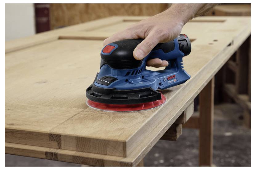 Een persoon gebruikt een draagbare elektrische schuurmachine op een houten oppervlak, waarbij hij de rand van een lichtgekleurde houten plank glad maakt in een werkplaats.