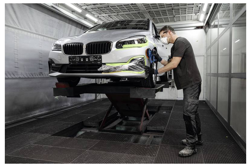 Een technicus in een spuitcabine poliert een witte auto op een hydraulische lift, waarbij hij zorgt voor een gladde afwerking. De omgeving is goed verlicht en industrieel.