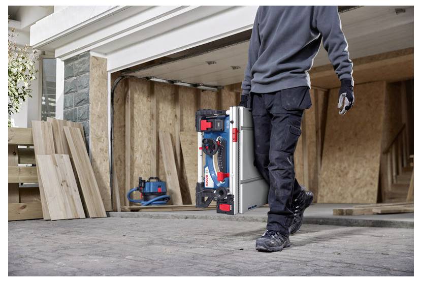Een persoon draagt een draagbare tafelzaag buiten, in de buurt van een garage in aanbouw met multiplex wanden en verspreid liggende houten planken.
