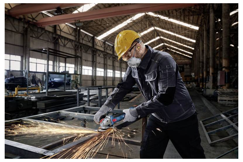 Een arbeider in veiligheidsuitrusting gebruikt een haakse slijper op metaal, waarbij vonken ontstaan, in een grote industriële loods met hoge plafonds.