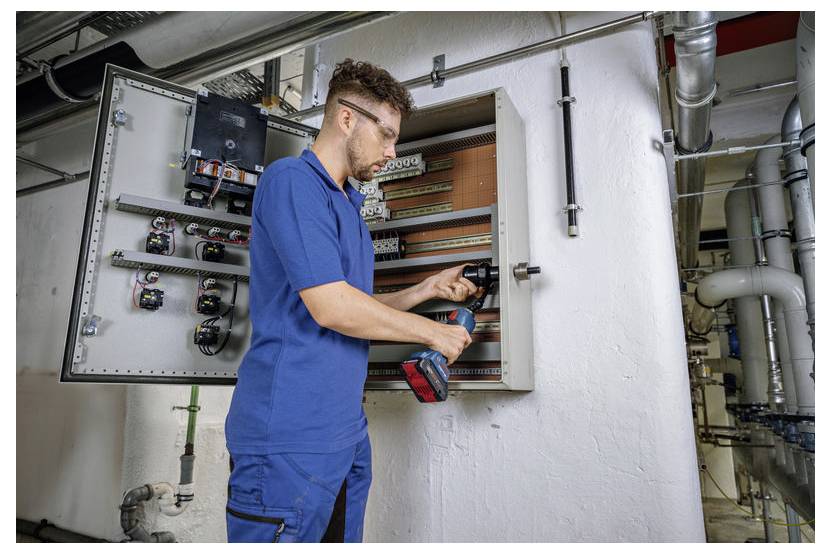Een persoon in een blauwe werkkleding die een boormachine gebruikt om apparatuur te repareren of te installeren in een open elektrisch paneel in een industriële omgeving.