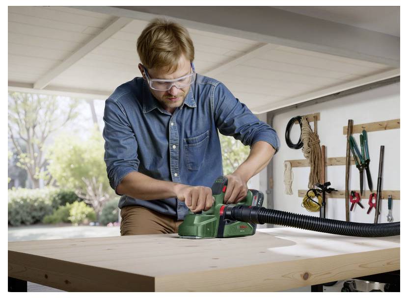 Een persoon die een elektrische schuurmachine gebruikt op een houten tafel in een werkplaats, met veiligheidsbril. Tuin is zichtbaar door het raam. Gereedschappen hangen aan de muur.