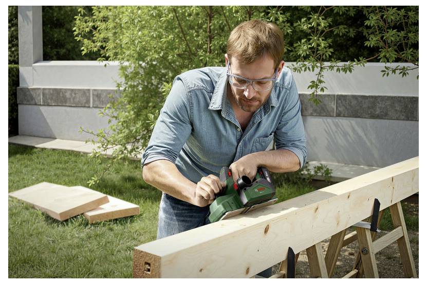 Een man met veiligheidsbril gebruikt een elektrisch gereedschap om een houten balk glad te schuren in de buitenlucht, met meer hout en groen op de achtergrond.