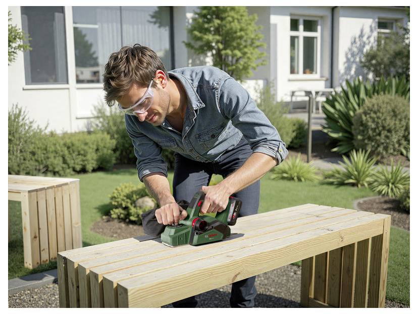 Een persoon met veiligheidsuitrusting gebruikt een elektrisch gereedschap om een houten bank te schaven in de openlucht, omringd door planten en met een gebouw op de achtergrond.