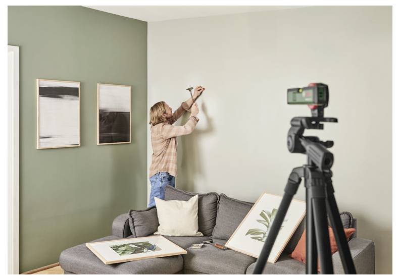 Een persoon op een bank hamert een spijker in de muur om foto's op te hangen, terwijl een camera op een statief het proces vastlegt.