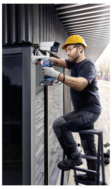 Een werknemer met een gele veiligheidshelm installeert een beveiligingscamera op de buitenkant van een gebouw, waarbij hij een boormachine gebruikt terwijl hij op een ladder staat.