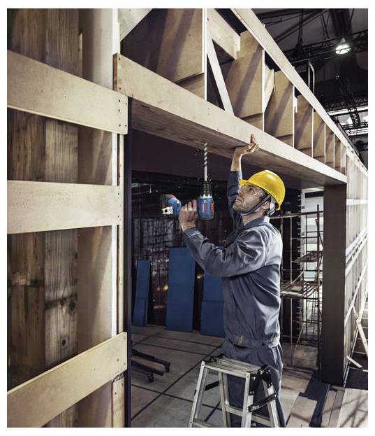 Een bouwvakker met een gele veiligheidshelm staat op een ladder en boort in een houten balk in een grote industriële constructie.