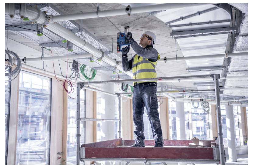 Een bouwvakker op een steiger boort in een plafond, omringd door gereedschappen en draden in een gedeeltelijk gebouwde industriële ruimte.