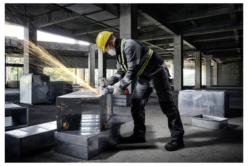 Een bouwvakker in veiligheidsuitrusting slijpt een metalen kanaal, waarbij heldere vonken vrijkomen, in een onafgewerkt gebouw met blootgestelde betonnen balken.
