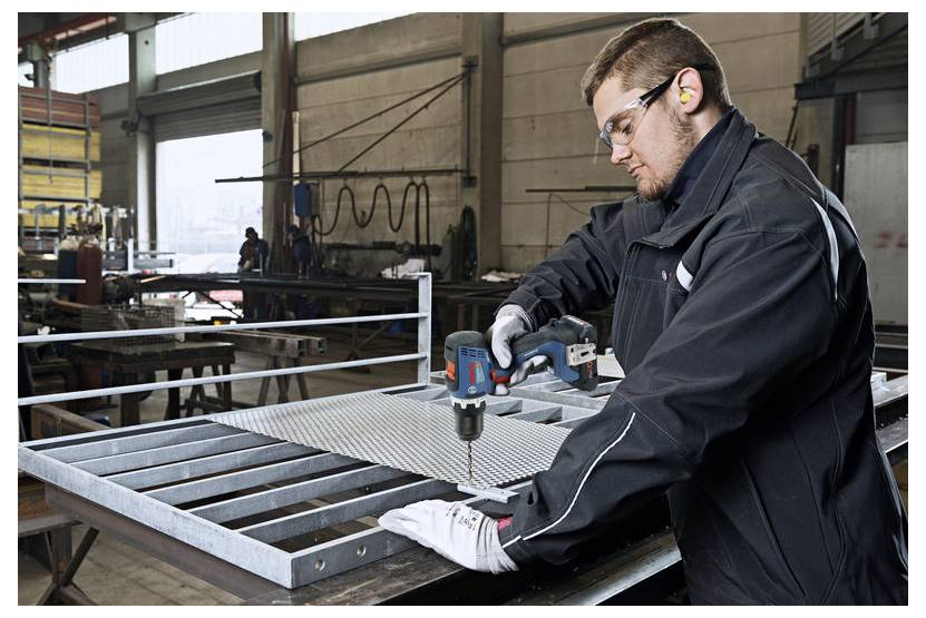 Een werknemer met veiligheidsuitrusting bedient een elektrisch gereedschap om metaal te boren in een industriële werkplaats.