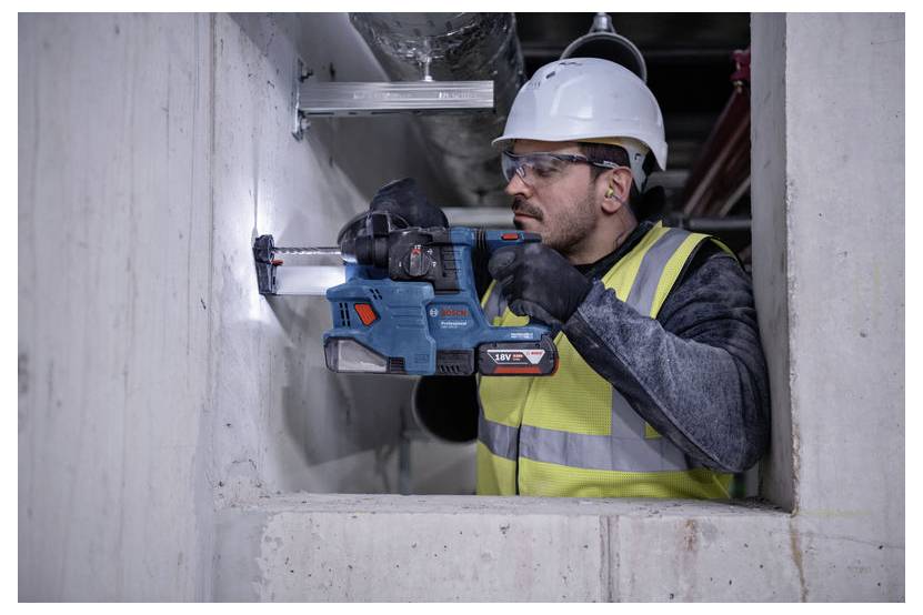 Een bouwvakker met een veiligheidshelm, veiligheidsbril en een reflecterend veiligheidshesje boort met een accuboormachine in een betonnen muur.