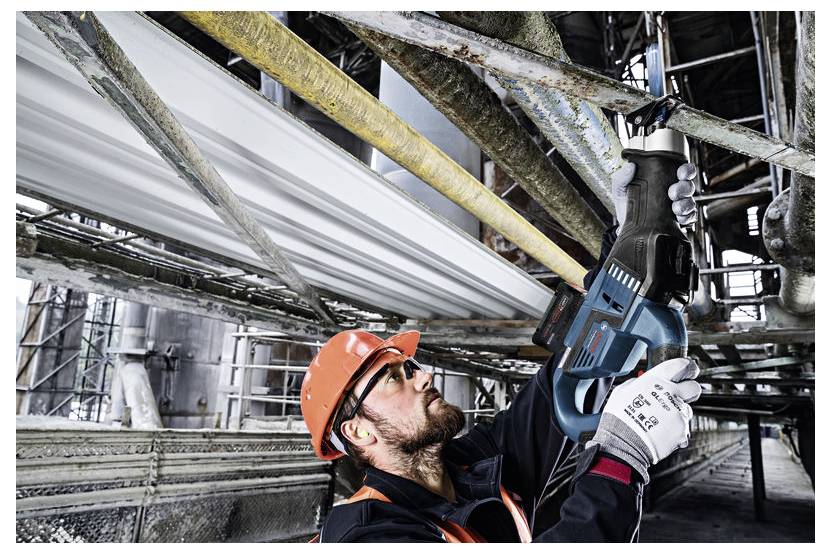 Een bouwvakker met een helm en veiligheidsuitrusting gebruikt een elektrische zaag om metalen buizen door te snijden op een industrieterrein.
