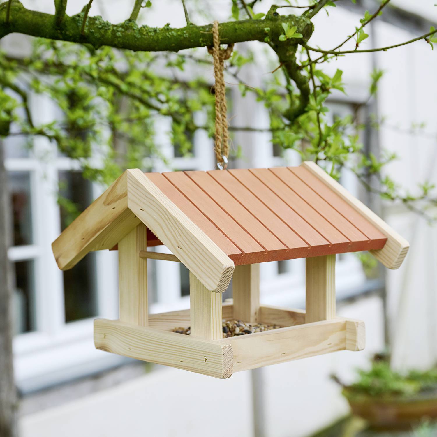 Een houten vogelvoeder met een rood dak hangt aan een boomtak, gevuld met zaden, in een tuinomgeving met een raam op de achtergrond.