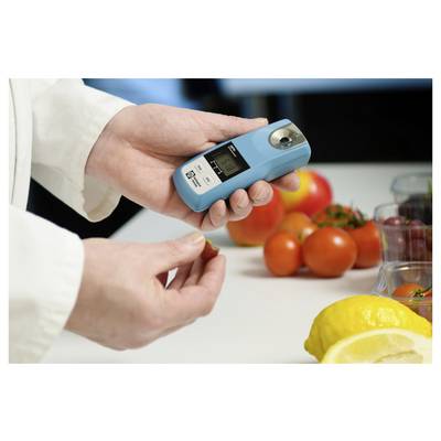 Een persoon gebruikt een digitale refractometer om suikergehaltes in fruit te meten. Tomaten en citroenen liggen op tafel, wat wijst op een laboratoriumomgeving.