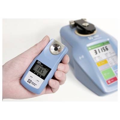 Een hand houdt een digitale refractometer vast die '38,5' meet. Een grotere blauwe meetapparatuur met een digitaal display bevindt zich op de achtergrond.