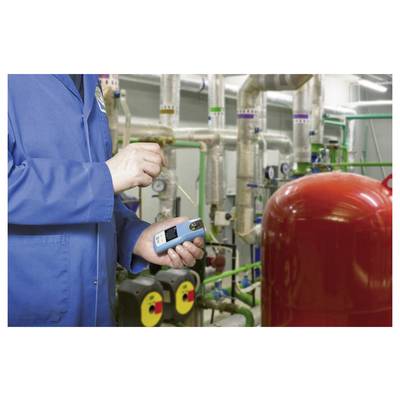 Een persoon in een blauwe laboratoriumjas gebruikt een draagbaar apparaat om gegevens te meten in een industriële omgeving met buizen en machines.