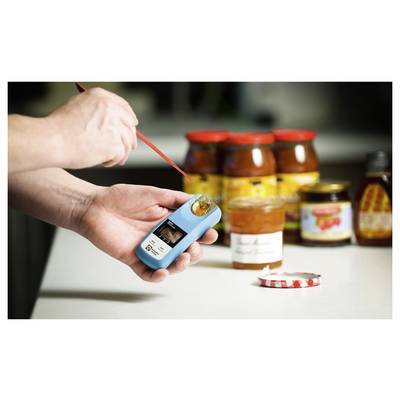 Handen die honing testen met een refractometer in een keuken, met potten honing en inmaakpotten op de achtergrond.