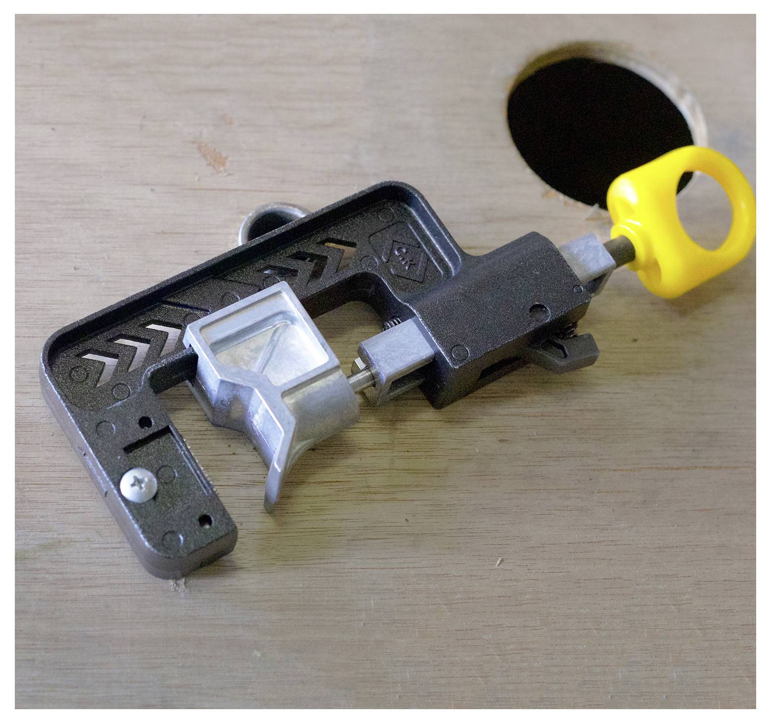 Een gereedschap voor houtbewerking staat op een houten oppervlak, gedeeltelijk uitstekend boven een gat. Het gereedschap heeft een gele handgreep en een metalen klem.