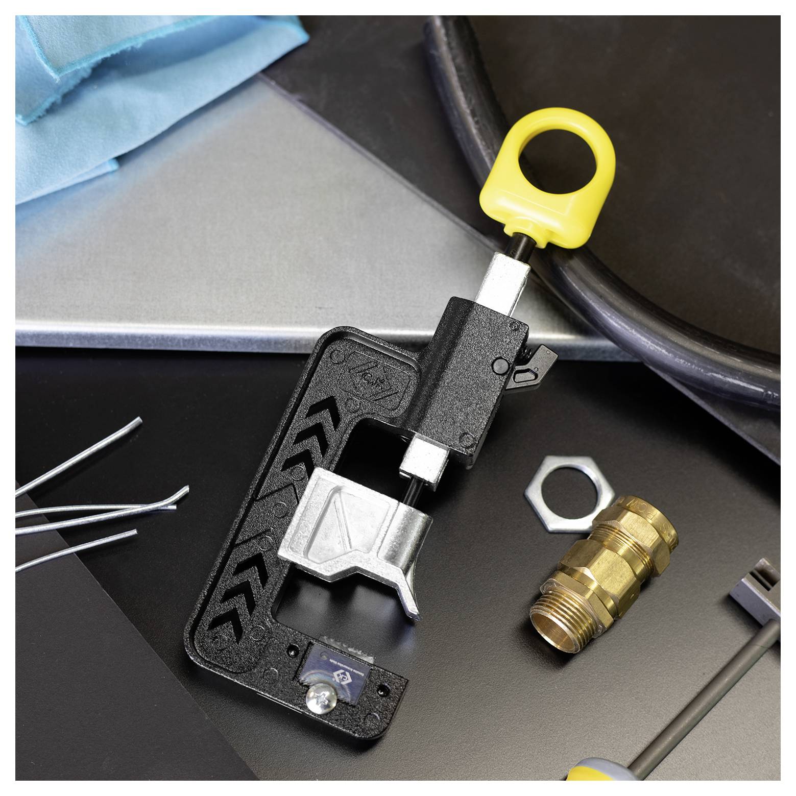 Een metalen pijpsnijder met een gele handgreep staat op een oppervlak met verschillende gereedschappen en een zeskantmoer in de buurt.