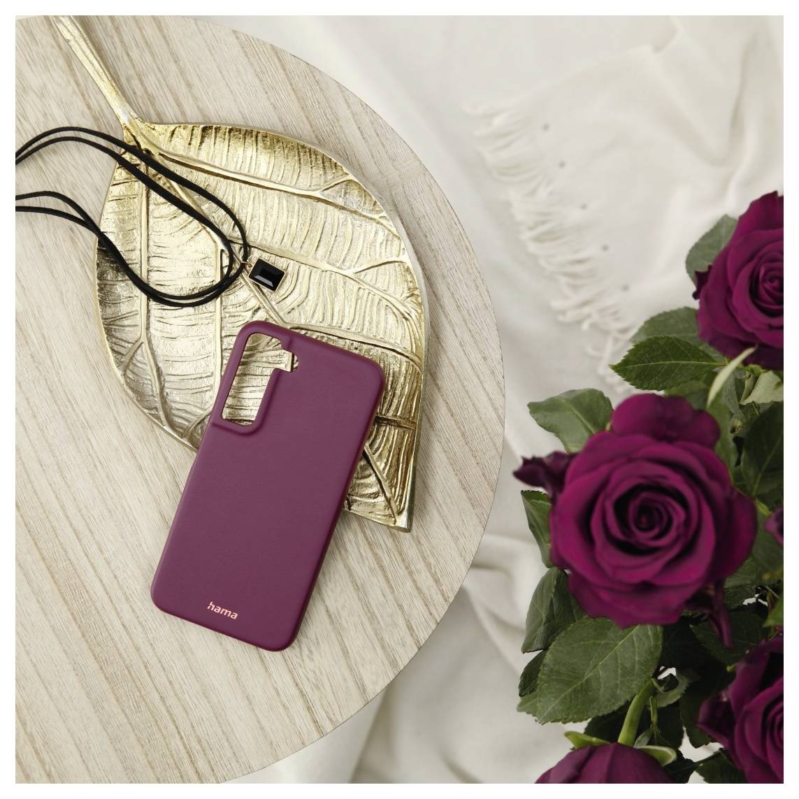 Een telefoonhoesje in bordeauxrood op een tafel naast een gouden bladornamement, een zwart snoer en een boeket donkerpaarse rozen.