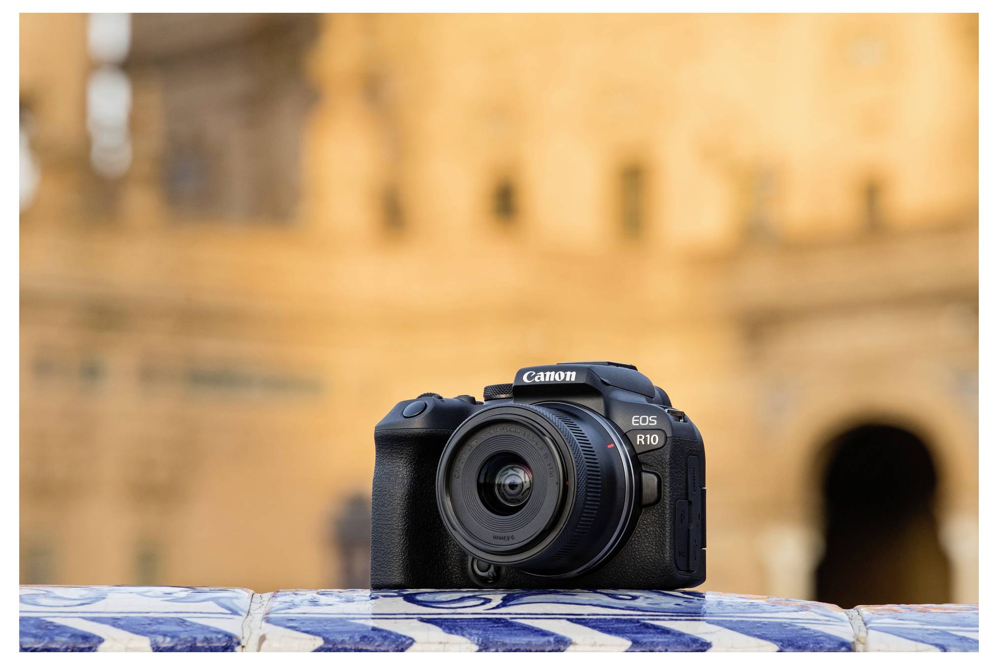 Een Canon EOS R10-camera die rust op een betegelde rand met wazig historische architectuur op de achtergrond.