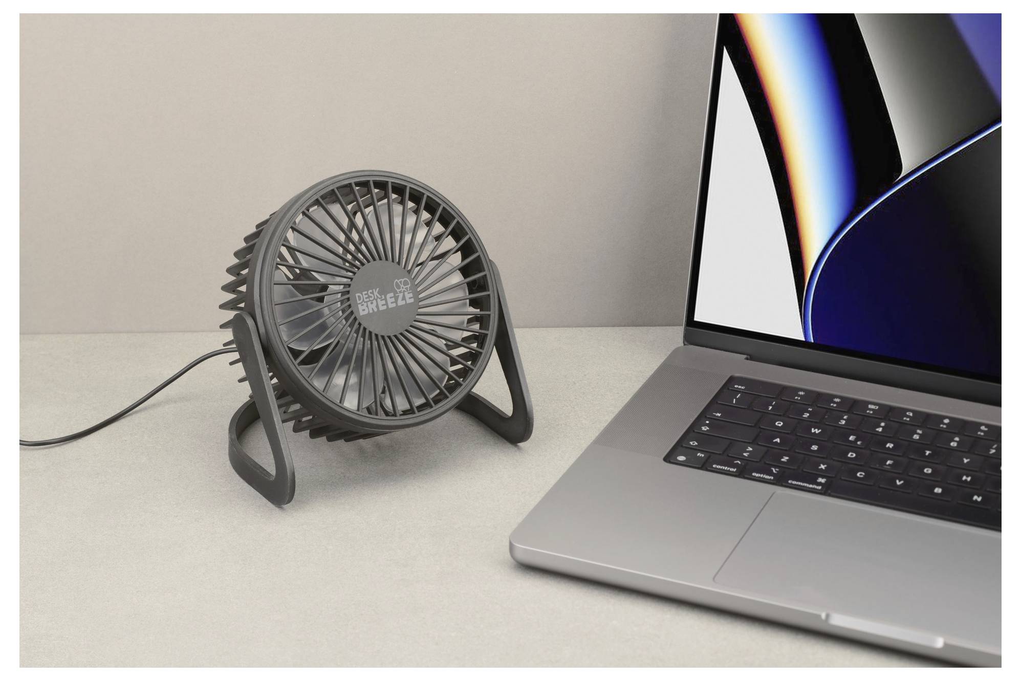 Een kleine bureauventilator aangesloten op een laptop, staande op een licht gekleurde ondergrond. Het laptopscherm is aan en toont een donker abstract ontwerp.