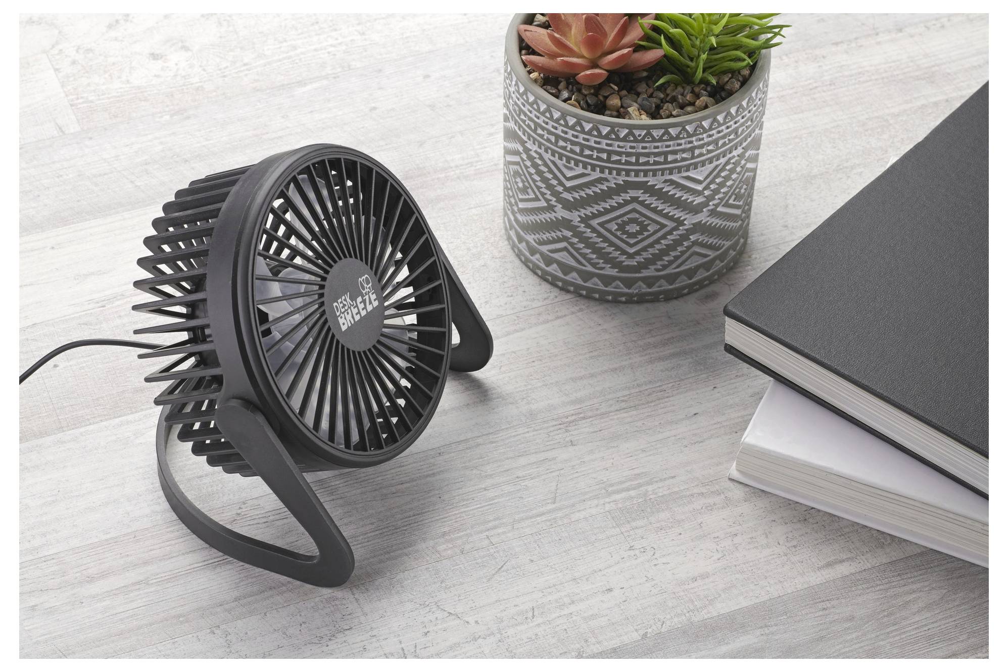 Een kleine bureauventilator staat op een houten oppervlak naast een gemaakt potje met vetplanten en gestapelde notitieboeken.