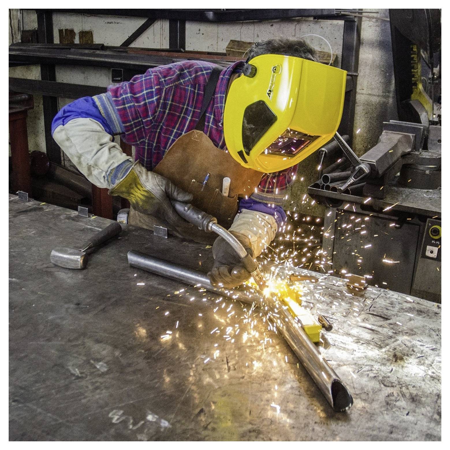 Een persoon met een lasmasker en beschermende uitrusting is metalen onderdelen aan het lassen op een werkbank, waarbij heldere vonken ontstaan in een industriële omgeving.