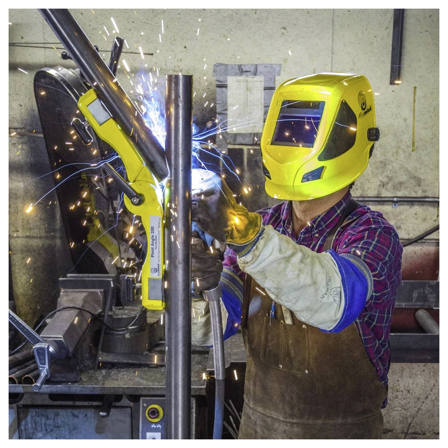 Een persoon met een gele lashelm en beschermende uitrusting last een metalen pijp, waarbij vonken zichtbaar zijn in een werkplaatsomgeving.
