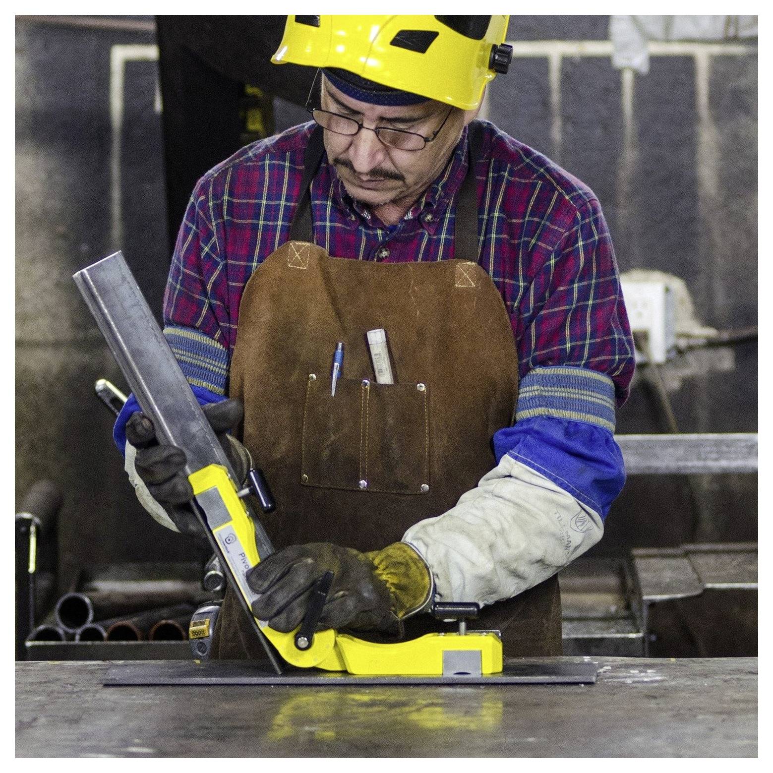 Een persoon in veiligheidsuitrusting, inclusief een gele helm en bruine schort, gebruikt een meetinstrument om de uitlijning te controleren op een metalen onderdeel in een werkplaats.