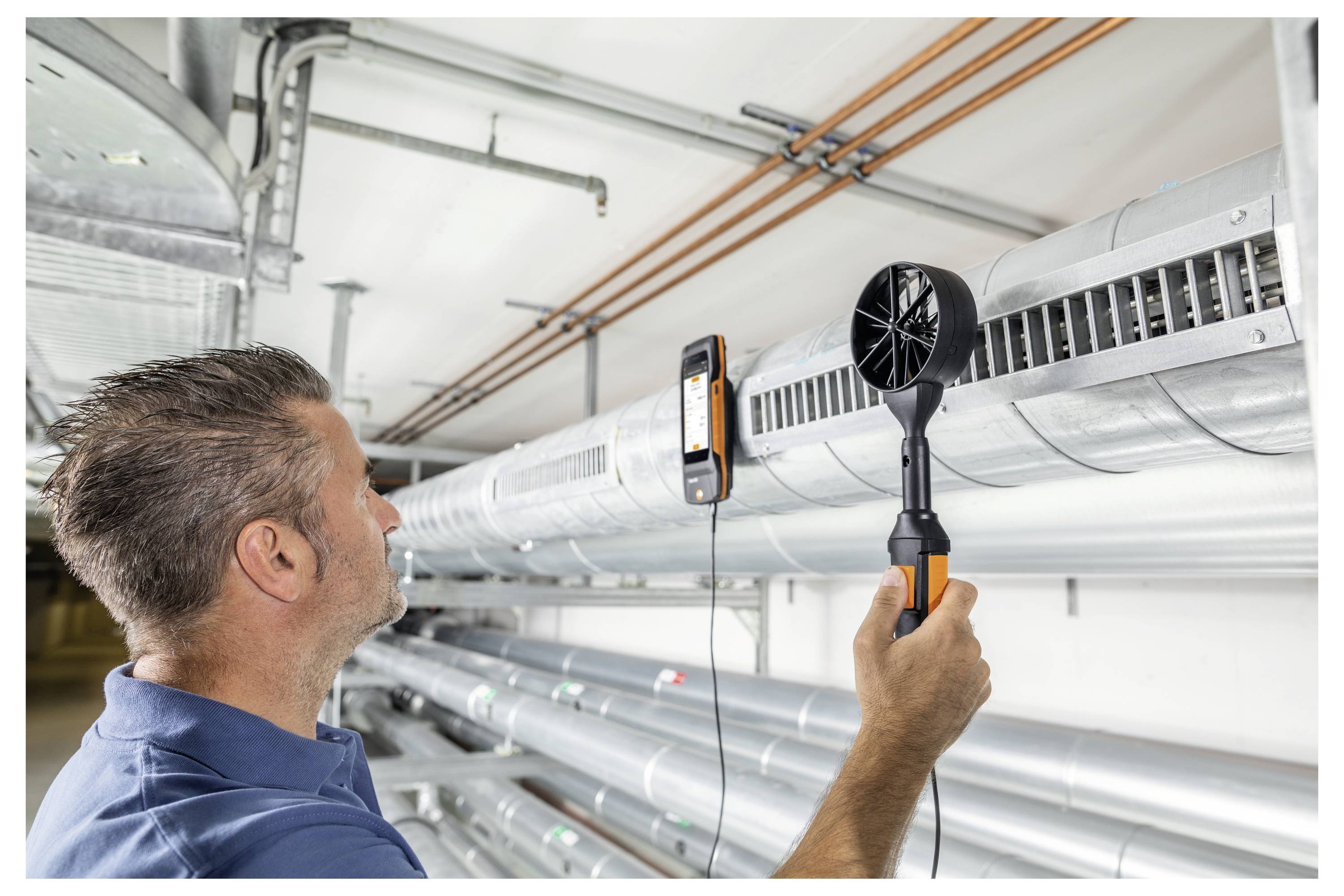 Een man in een blauw shirt gebruikt een handanemometer om de luchtstroom te meten vanuit een grote metalen ventilatiebuis in de technische ruimte van een gebouw.