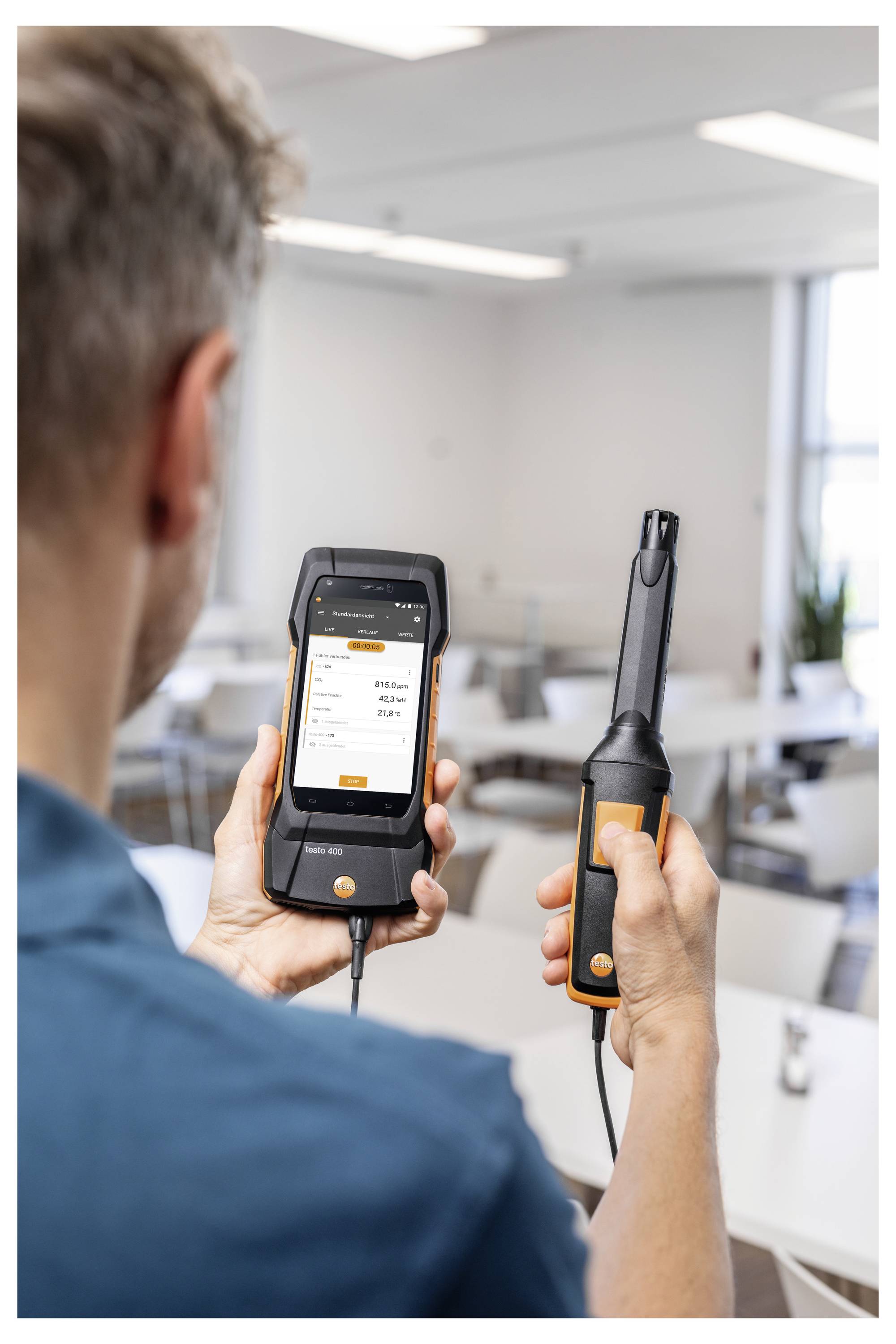 Een persoon houdt een luchtkwaliteitsmeetapparaat en een mobiel apparaat vast waarop luchtkwaliteitsgegevens op het scherm worden weergegeven, in een moderne kantoorruimte.