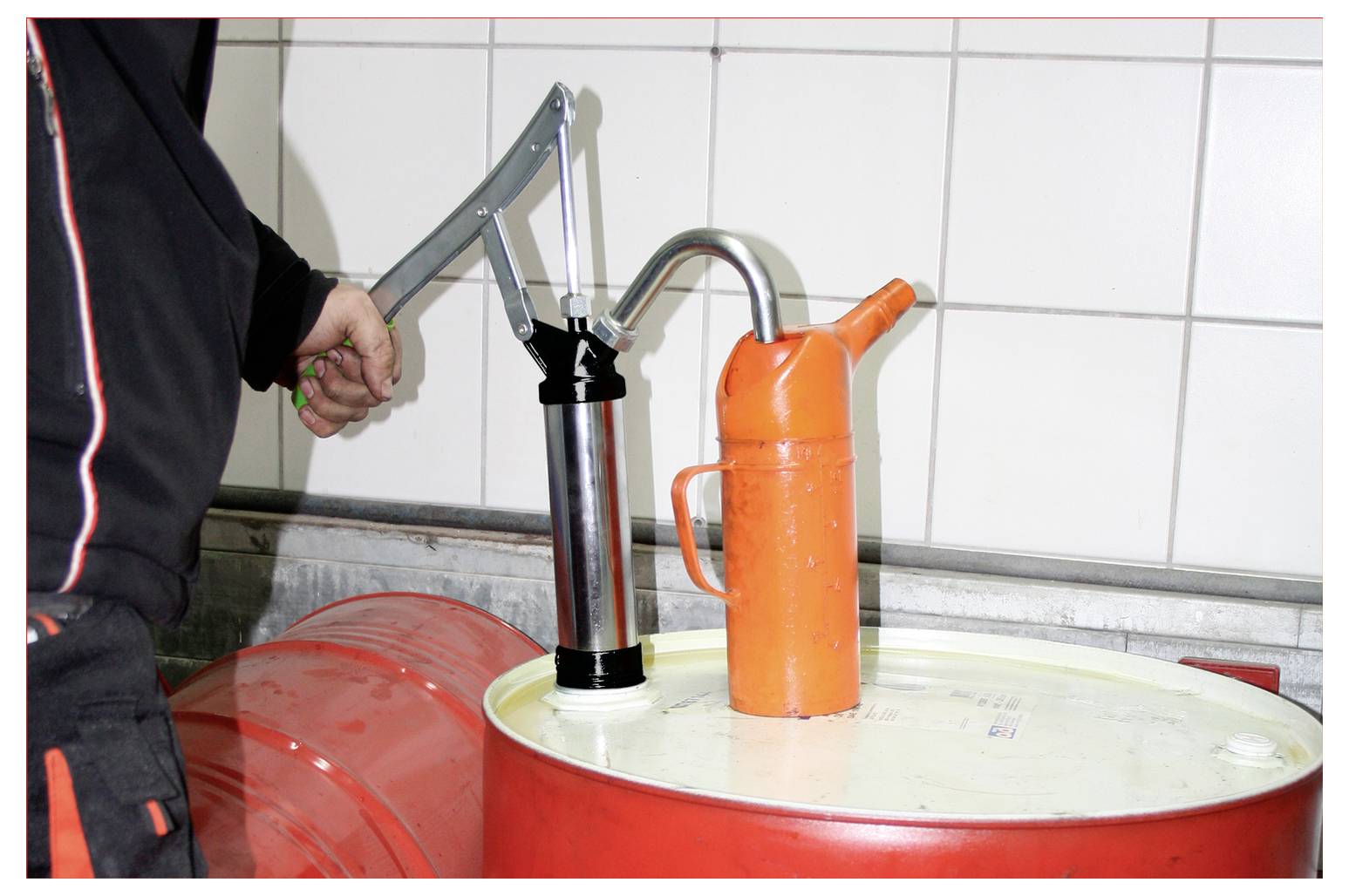 Een persoon bedient een handmatige pomp die is bevestigd aan een grote rode vat, waarbij vloeistof wordt afgegeven in een oranje container bovenop het vat.