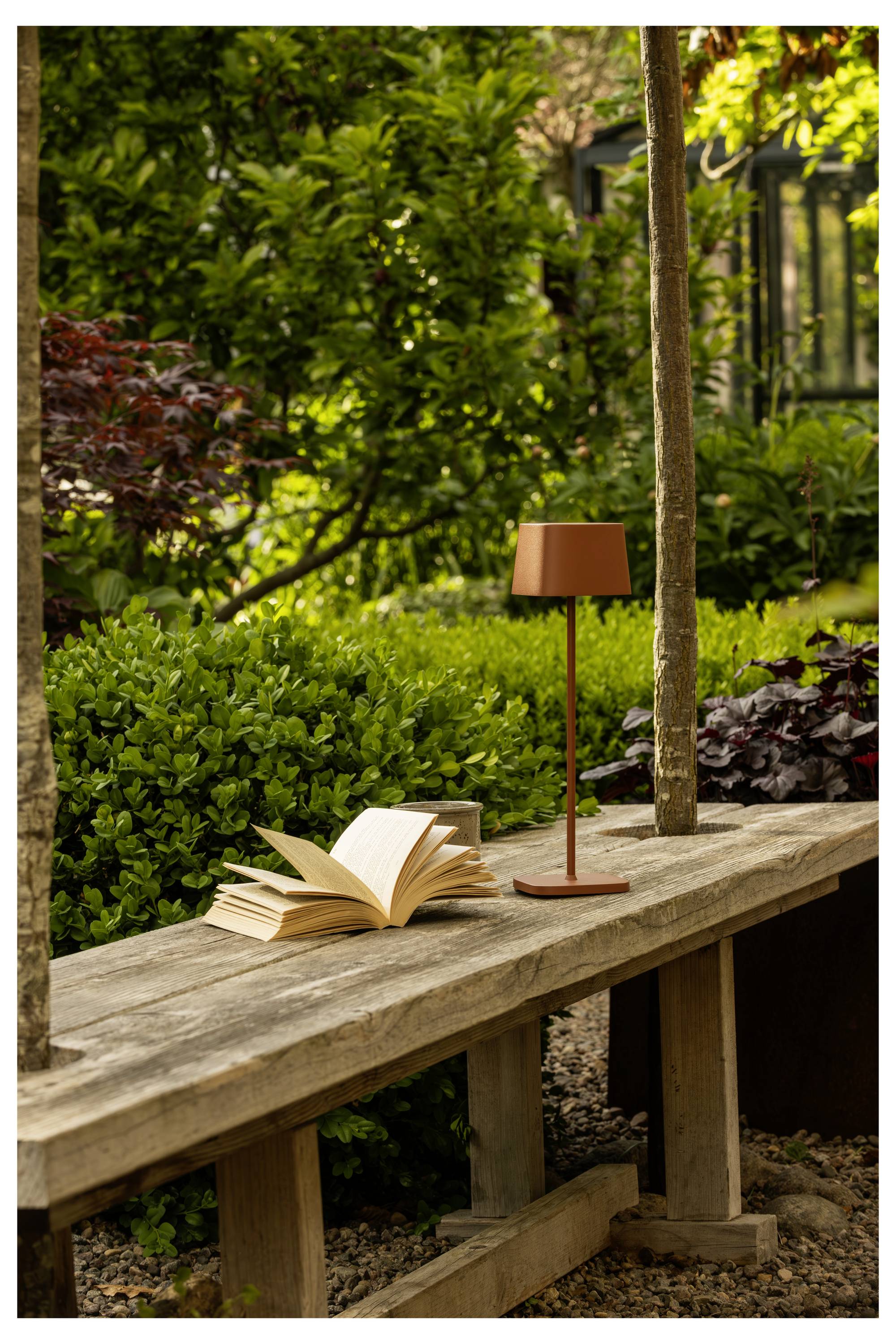 Een houten bank in een tuin met een opengeslagen boek en een kleine lamp erop, omringd door weelderig groen gebladerte en zacht natuurlijk licht.