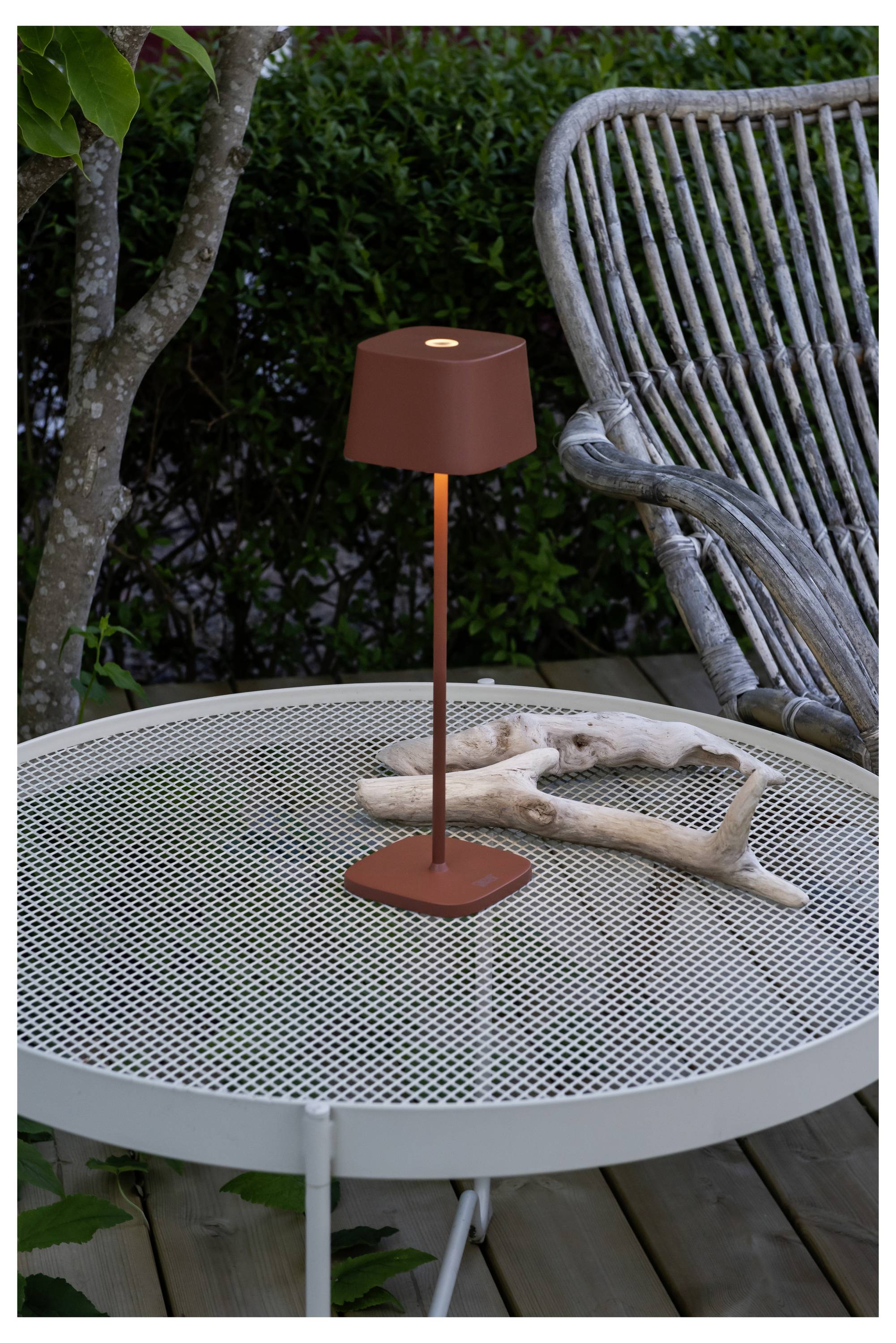 Een kleine, rode buitenlamp op een witte metalen tafel naast decoratief drijfhout; een rieten stoel en groen zijn op de achtergrond.