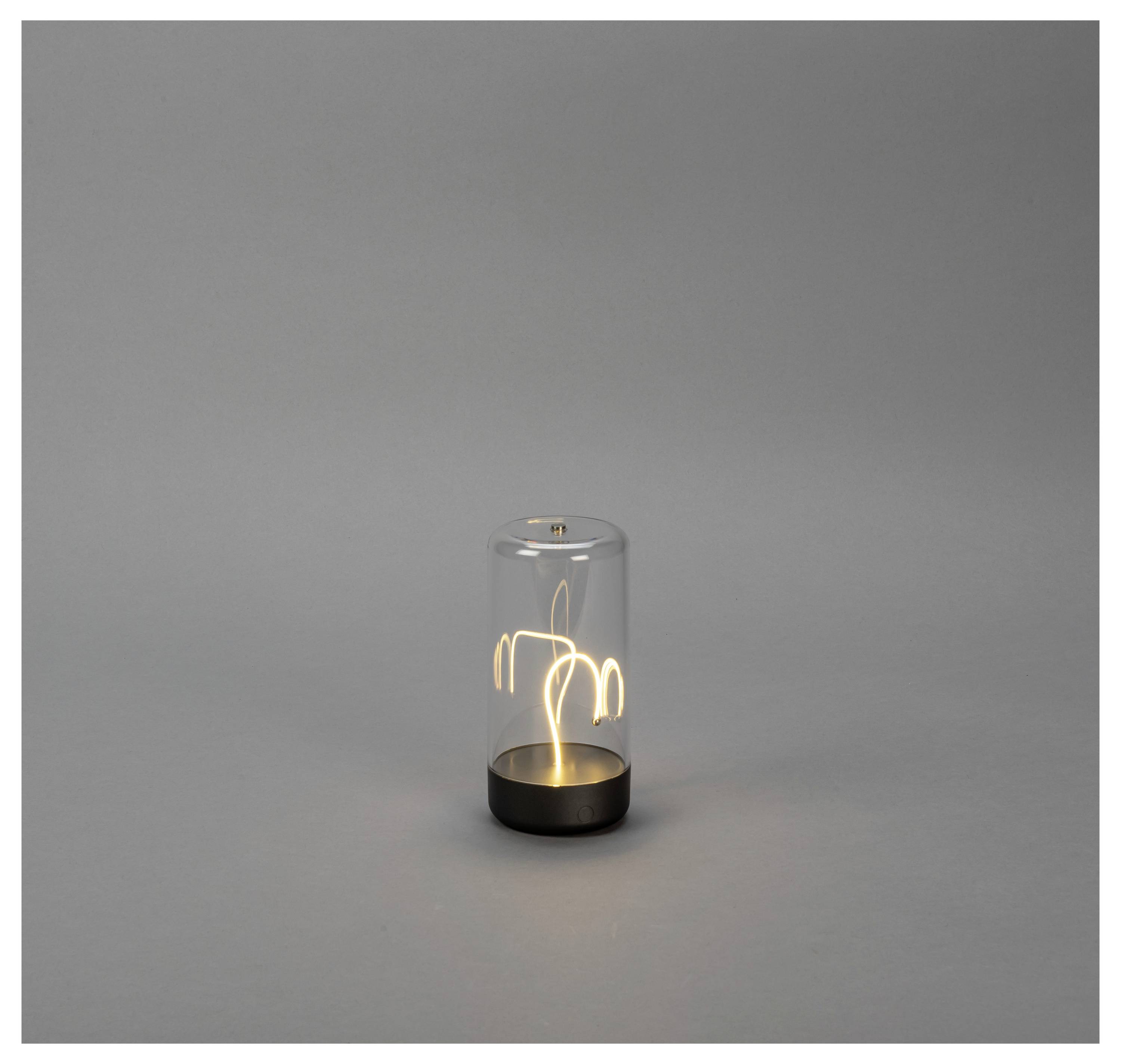 Een kleine, transparante glazen lamp met een zwarte voet geeft zacht licht, waarbij een abstract gloeidraadontwerp binnenin wordt getoond. Hij staat tegen een grijze achtergrond.