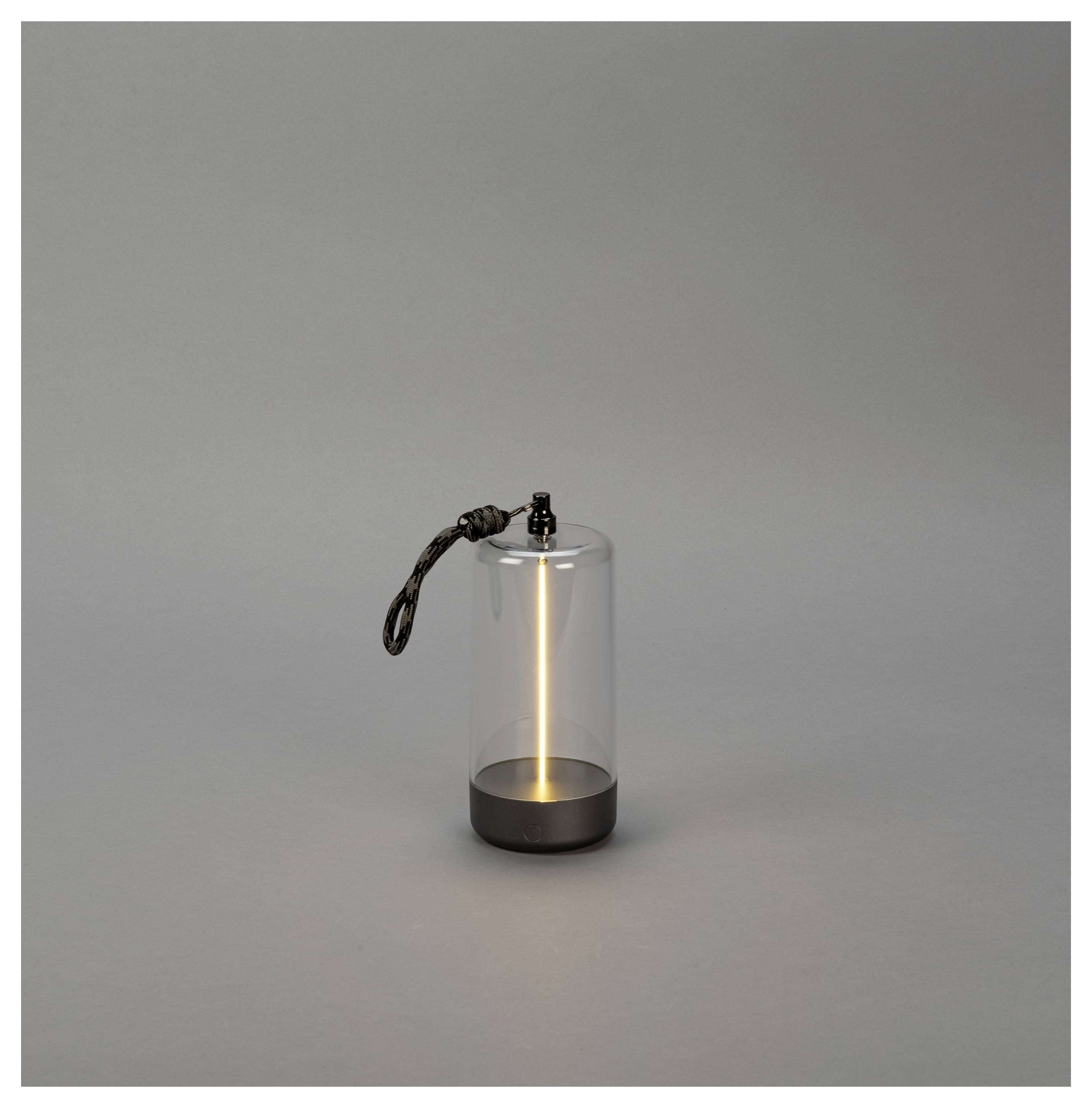 Een cilindrische lantaarn met een gloeiend licht binnenin staat op een vlak, grijs oppervlak. Hij heeft een touwgreep die aan de bovenkant is bevestigd.