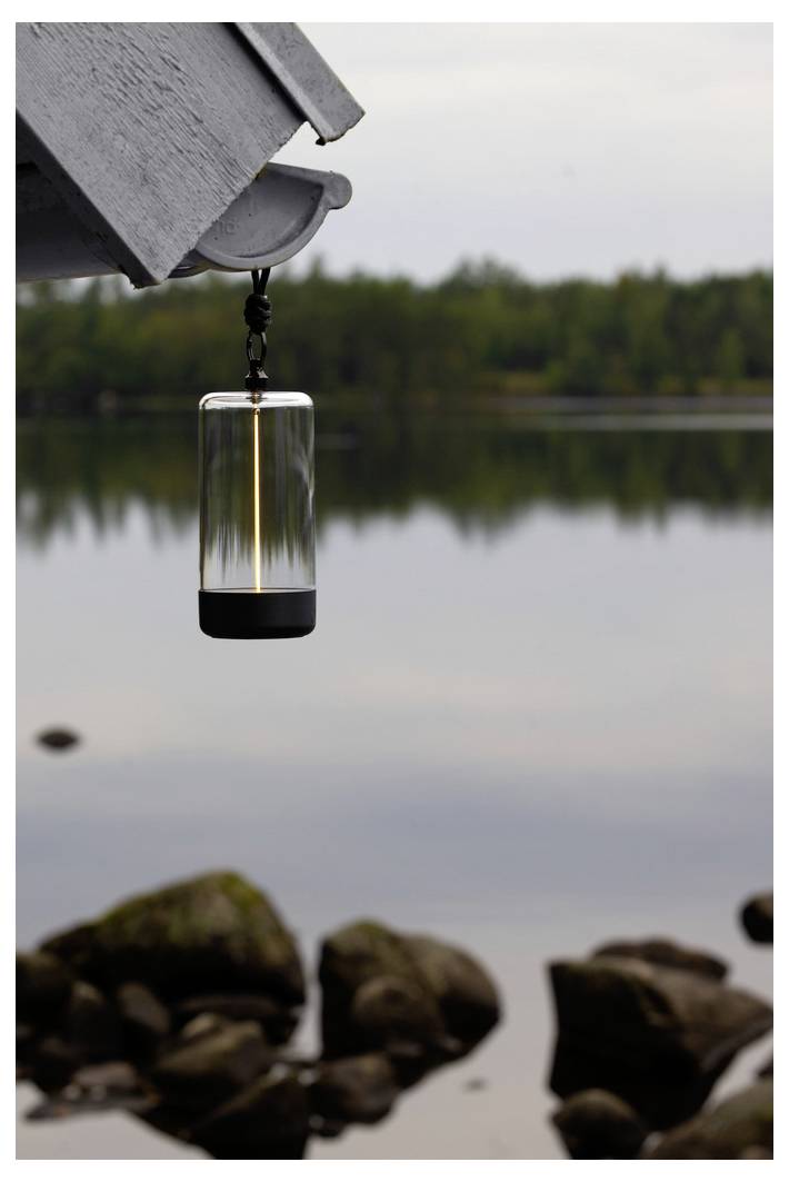 Een lantaarn hangt aan een houten constructie bij een rustig meer, met rotsen op de voorgrond en een bebost meer op de achtergrond.