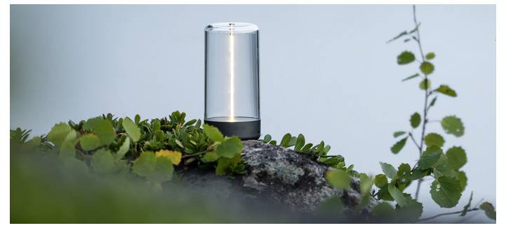 Een kleine, cilindrische buitenlamp staat op een rots omgeven door groen gebladerte, waarbij hij een zacht licht uitstraalt tegen een wazig, licht achtergrond.