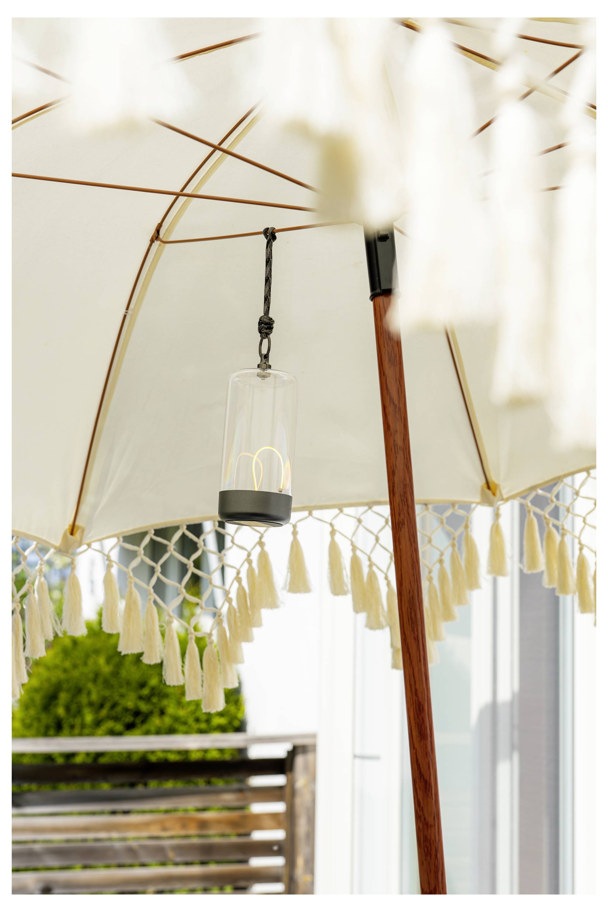 Een witte buitenparasol met franjes beschermt een tuingedeelte tegen de zon. Een kleine cilindrische glazen lantaarn hangt aan de parasol, waardoor een decoratief accent wordt toegevoegd.