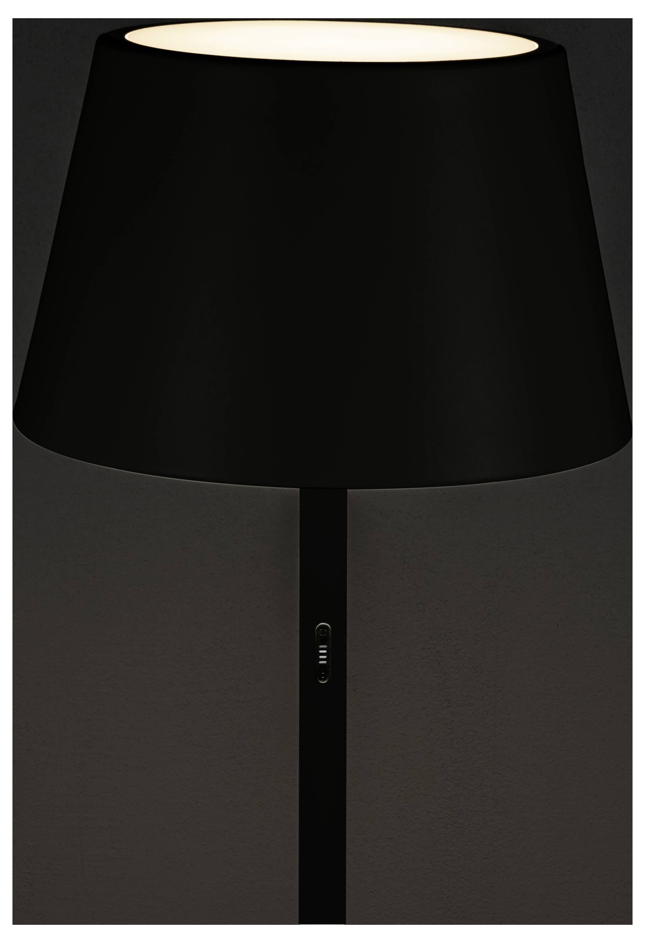 Een moderne zwarte tafellamp met een brede cilindrische kap die een donkere kamer zacht verlicht.