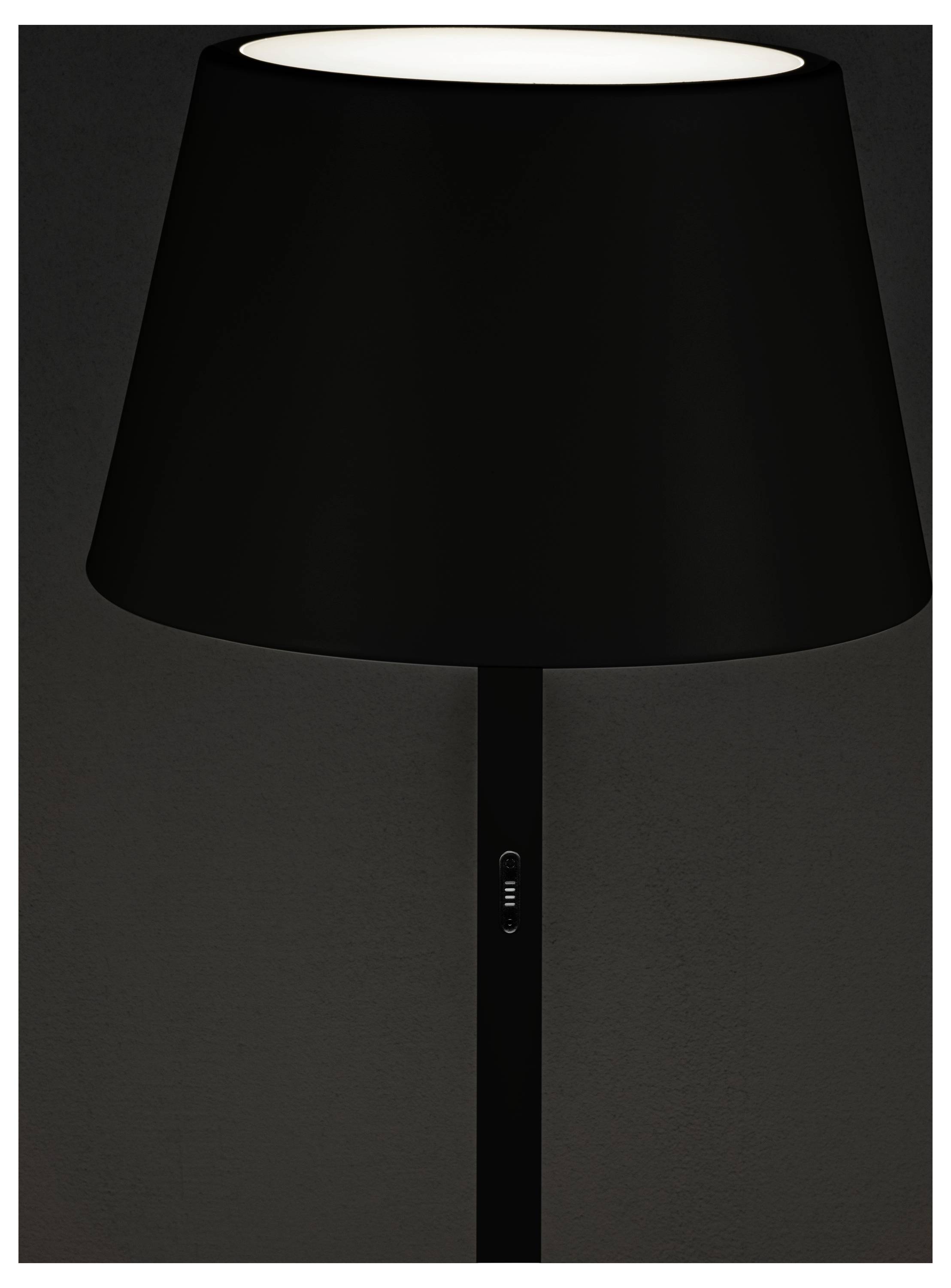 Een zwarte tafellamp met een grote kap is verlicht tegen een donkere achtergrond, waarbij het moderne en minimalistische ontwerp wordt benadrukt.