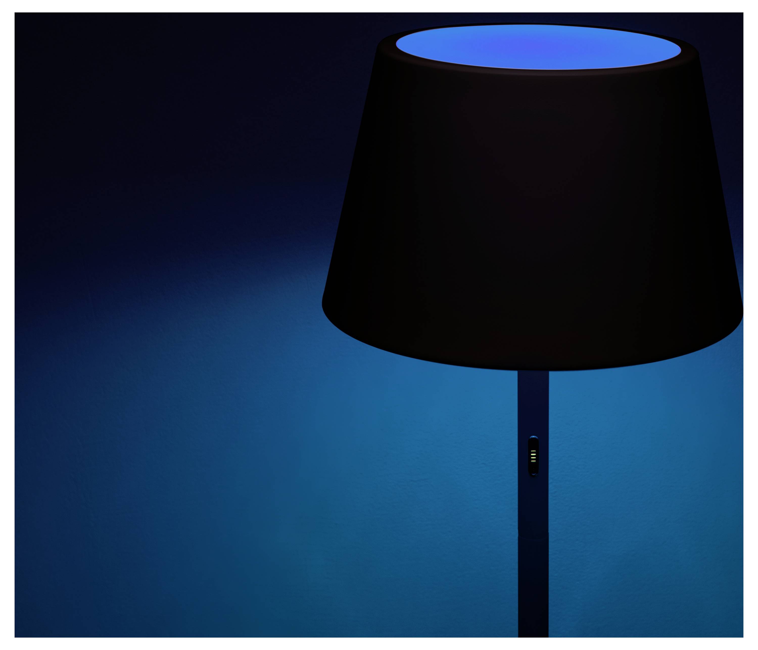 Een zwarte lampenkap met een oplichtend blauw licht bovenaan staat in een donkere kamer, waarbij een subtiele blauwe gloed op de donkere muur wordt geworpen.